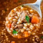 A close-up of a ladle holding homemade beef barley soup, full of hearty chunks of beef, barley, carrots, and herbs in a rich brown broth. The ladle hovers above the pot, capturing the soup's texture and ingredients beautifully.