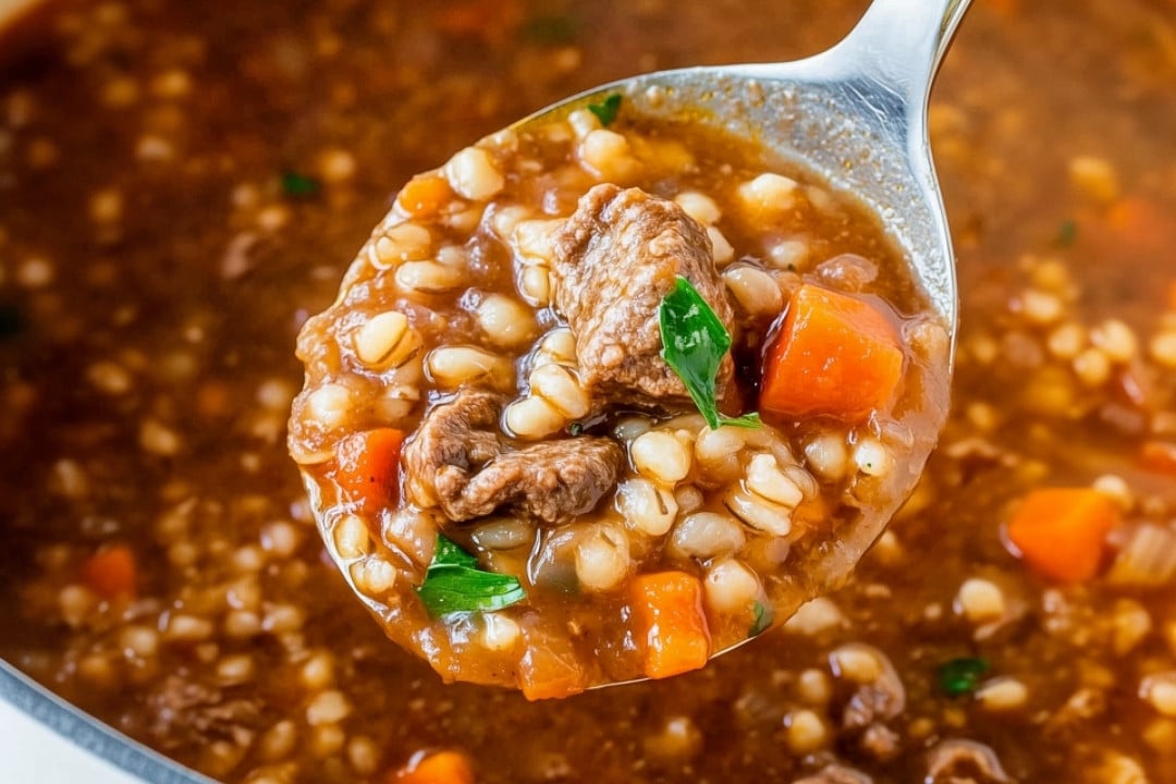 A close-up of a ladle holding homemade beef barley soup, full of hearty chunks of beef, barley, carrots, and herbs in a rich brown broth. The ladle hovers above the pot, capturing the soup's texture and ingredients beautifully.