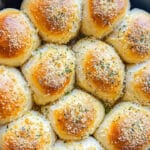 A black pan filled with freshly baked golden-brown dinner rolls, reminiscent of cheesy Buffalo chicken bombs, is topped with garlic, herbs, and grated cheese. The rolls are arranged in a circular pattern, creating an appetizing, rustic presentation.