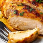 Close-up of a juicy, oven-baked chicken breast sliced and garnished with fresh herbs. A fork rests on the plate, accentuating the golden-brown texture of this easy baked chicken breast.