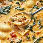 A close-up of a creamy chicken soup with pasta featuring shell pasta, shredded chicken, and fresh spinach, garnished with red pepper flakes. A spoon is lifting a portion, showcasing the hearty ingredients in a rich broth reminiscent of a Marry Me Chicken Soup recipe.