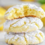 Three chewy lemon cookies are stacked, with the top one broken in half to reveal a soft interior. The easy lemon crinkle cookies are coated in powdered sugar, and fresh lemons are blurred in the background.