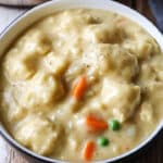 A bowl of easy, creamy chicken and dumplings soup, featuring fluffy dumplings, diced carrots, and green peas, topped with a sprinkle of black pepper for a comforting one-pot meal.