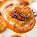 A fork holds a sweet potato slice topped with toasted marshmallow, brown sugar, and a pecan. Other similar sweet potato bites with marshmallows are blurred in the background on a white plate—a perfect holiday sweet potato side dish.