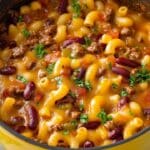 A pot filled with homemade chili mac, blending cheesy macaroni pasta with ground beef, kidney beans, and tomatoes, garnished with fresh parsley. The hearty dish is flavorful and comforting, crowned with a golden cheese topping.