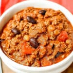 A bowl of hearty pumpkin chili with ground beef, kidney beans, and red bell peppers in a rich tomato sauce. A silver spoon is placed on the right, and a red cloth napkin is partially visible in the background. Perfect for anyone searching for an easy pumpkin chili recipe.