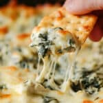 A hand dips a cracker into a creamy spinach and artichoke dip, with melted cheese stretching from the dish. The best spinach artichoke dip recipe delivers a delightful mix of visible spinach and artichokes under a golden-brown, bubbly top.