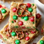 A stack of easy Christmas cookie bars topped with red and green candies and pretzels. The festive bars rest invitingly on a light-colored surface, accompanied by a steaming cup of hot chocolate.