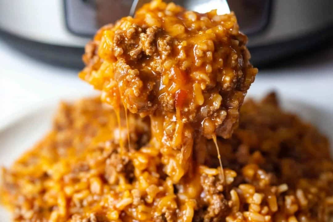 A spoonful of cheesy taco beef and rice is being lifted from a plate, with strings of melted cheese extending decadently. The dish appears hearty and savory, with a slow cooker partially visible in the background, hinting at its effortless preparation.