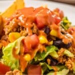 A close-up of an easy taco salad showcases crispy lettuce, diced tomatoes, black beans, corn, shredded cheese, and seasoned ground beef. The salad is elegantly topped with a generous drizzle of Catalina dressing, while crunchy taco shells linger invitingly in the background.