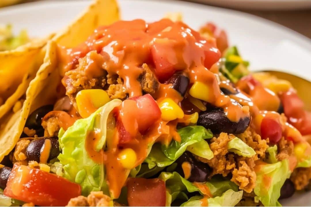 A close-up of an easy taco salad showcases crispy lettuce, diced tomatoes, black beans, corn, shredded cheese, and seasoned ground beef. The salad is elegantly topped with a generous drizzle of Catalina dressing, while crunchy taco shells linger invitingly in the background.