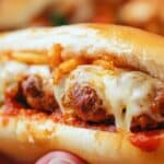 A close-up of an oven-baked meatball sub with three meatballs smothered in melted cheese inside a soft bread roll. The sandwich is topped with crispy onions and rests on a luscious layer of tomato sauce, perfect for anyone looking for an irresistible oven-baked meatball sandwiches recipe.