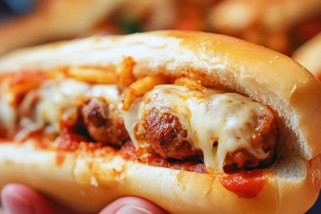 A close-up of an oven-baked meatball sub with three meatballs smothered in melted cheese inside a soft bread roll. The sandwich is topped with crispy onions and rests on a luscious layer of tomato sauce, perfect for anyone looking for an irresistible oven-baked meatball sandwiches recipe.