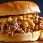 A root beer pulled pork sandwich topped with creamy macaroni and cheese, layered between a golden toasted bun. The sandwich sits on a wooden surface, with blurred macaroni in the background.