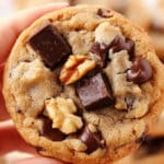 A hand holds a chocolate chip cookie with chunks of chocolate and walnut pieces, reminiscent of an easy banana bread cookie recipe. The golden-brown treat boasts a soft texture, with more cookies blurred in the background.