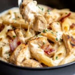 A bowl of creamy bacon chicken pasta featuring penne, grilled chicken, and bacon is garnished with herbs. A fork holds up a bite-sized portion, showcasing the rich Marry Me Chicken Pasta sauce and the dish's crispy texture.