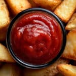 Close-up of a black bowl filled with homemade ketchup, surrounded by crispy potato wedges. The wedges are golden brown and appear freshly cooked, perfectly complementing the thick, red sauce in the center.