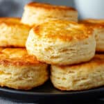 A stack of golden-brown biscuits piled high on a black plate. The biscuits have a slightly rough and uneven texture with visible flaky layers and a craggy top. They are stacked haphazardly, with some leaning against each other. The plate is sitting on a gray napkin.