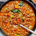 A bowl of easy lentil soup filled with carrots, celery, lentils, and greens, with a spoon gently resting in the bowl. The vegetarian soup recipe boasts a rich, broth-like appearance and is served elegantly on a striped cloth.