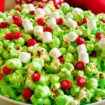 A bowl of holiday Grinch popcorn is topped with red candy-coated chocolates and small white marshmallows, creating a vibrant and festive snack.