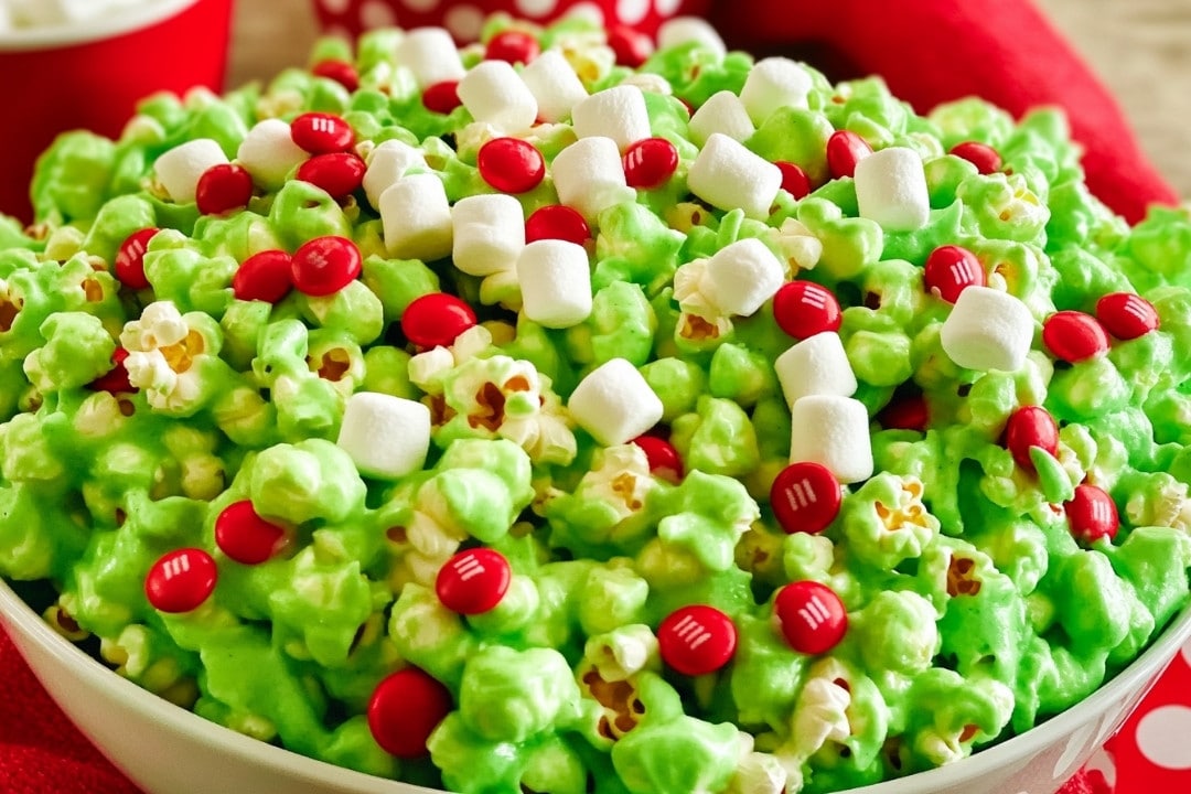 A bowl of holiday Grinch popcorn is topped with red candy-coated chocolates and small white marshmallows, creating a vibrant and festive snack.