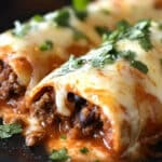 Close-up of two Beef & Bean Enchiladas on a black plate, filled with meat and topped with melted cheese and chopped cilantro. The tortillas are covered in a rich, savory sauce, making this an easy enchiladas recipe to savor.