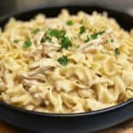 A black bowl filled with creamy Chicken & Noodles Crock Pot delight is topped with shredded chicken and garnished with fresh parsley. The dish rests on a wooden surface, highlighting its rich, savory texture.