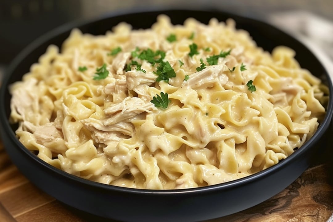 A black bowl filled with creamy Chicken & Noodles Crock Pot delight is topped with shredded chicken and garnished with fresh parsley. The dish rests on a wooden surface, highlighting its rich, savory texture.