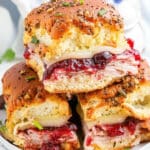 Three turkey and cranberry sliders stacked on a plate, featuring sliced turkey, melted cheese, and cranberry sauce nestled in poppy seed-topped Hawaiian rolls. This Cranberry Turkey Sliders recipe is garnished with chopped parsley and set against a white background with blueberries nearby.