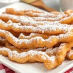 A plate of funnel cake fries dusted with powdered sugar sits on a red and white checkered tablecloth. The golden-brown treats are curled and crispy, with a jar of sugar and a wooden brush visible in the background.