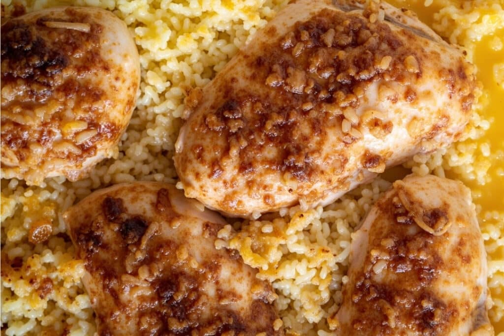 Close-up of a one-pan chicken and rice dish. The baked chicken breasts, seasoned with a golden-brown marinade, rest on fluffy rice. Garnished with minced garlic or onion, the crispy texture of this easy meal adds a delightful touch, making it perfect for any dinner table.