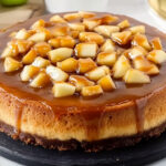 A caramel apple cheesecake sits on a black serving plate, topped with succulent caramel-coated apple chunks. The golden-brown glaze drips enticingly down the sides, exuding an irresistible charm. A spoon rests in the background, tempting you to dig into this easy cheesecake delight.