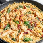 A skillet filled with creamy Tuscan chicken orzo, featuring slices of grilled chicken, sun-dried tomatoes, and fresh spinach. This easy pasta recipe is garnished with black pepper, making it warm and appetizing.