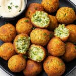 A black plate showcases a homemade falafel recipe with crispy golden-brown balls, some halved to reveal a green herb filling. Accompanied by a small dish of creamy white dipping sauce garnished with herbs, this presentation is both appetizing and inviting.