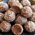 A black plate is stacked with round coconut energy balls, each sprinkled with shredded coconut. They are arranged neatly, creating a visually appealing display on a wooden table—a perfect healthy snack recipe for any time of the day.