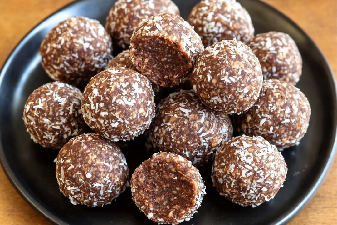 A black plate is stacked with round coconut energy balls, each sprinkled with shredded coconut. They are arranged neatly, creating a visually appealing display on a wooden table—a perfect healthy snack recipe for any time of the day.