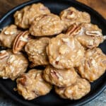 A black plate holds a stack of no-bake pecan coconut praline cookies, showcasing their light brown color and nutty texture with visible pecans. Nestled on a dark wooden board, the rustic presentation invites delight with this cozy cookie recipe.