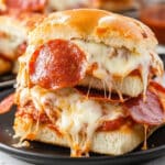 These cheesy pepperoni pizza sliders, with melted mozzarella and tomato sauce, are stacked elegantly on a small plate, showcasing tempting layers of bread and toppings. Blurred in the background is a cup of sauce. This party appetizer recipe is perfect for any casual gathering.