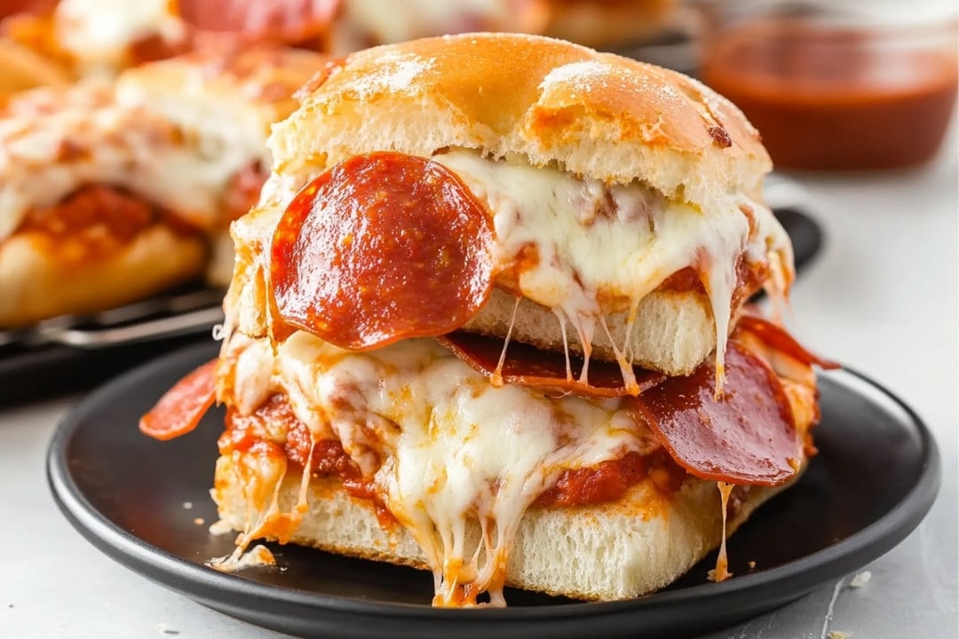 These cheesy pepperoni pizza sliders, with melted mozzarella and tomato sauce, are stacked elegantly on a small plate, showcasing tempting layers of bread and toppings. Blurred in the background is a cup of sauce. This party appetizer recipe is perfect for any casual gathering.