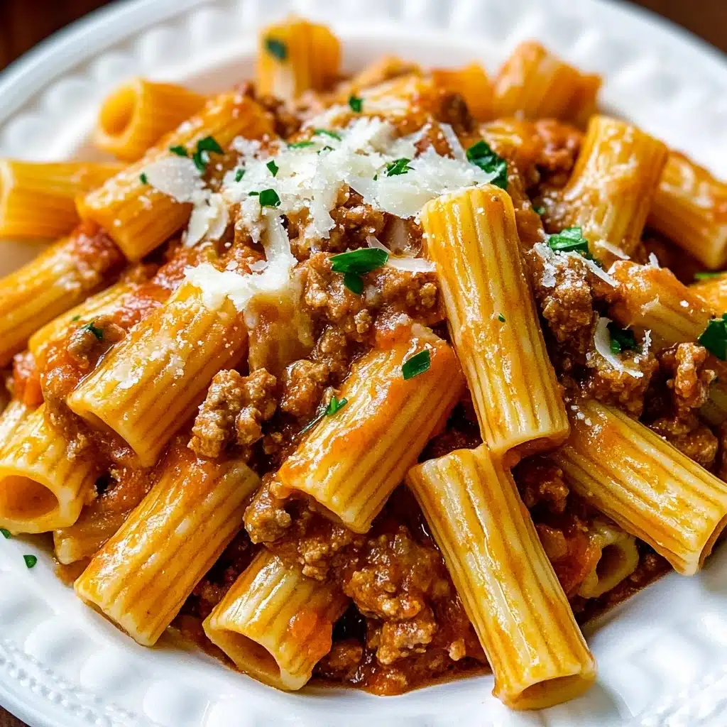 Italian Sausage Rigatoni with Spicy Cream Sauce, spicy sausage pasta, creamy rigatoni recipes, quick Italian pasta dishes, hearty sausage pasta A white plate is filled with large, ribbed rigatoni pasta, coated in a thick, creamy orange-red tomato sauce with bits of browned ground meat mixed in. The sauce clings to each tube of pasta and is generously scattered with small shreds of white cheese and a few pieces of fresh chopped green parsley on top. The pasta sits on a white marbled texture background, and everything looks glossy and rich, with some sauce pooling at the bottom.