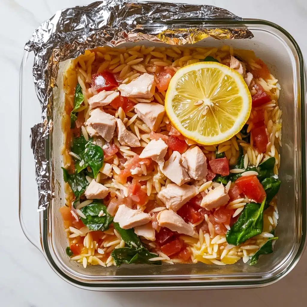 Greek Chicken Casserole, Greek Chicken Casserole recipe, Mediterranean chicken bake, feta chicken casserole, easy Greek chicken dinner A glass baking dish shows a colorful layered meal, with the bottom made of golden yellow orzo and bits of green vegetables, all slightly mixed but forming the base. On top of this, there are chunks of cooked chicken that are juicy and pale with a gentle texture. Scattered over everything are white crumbles of cheese, adding a little rough texture. Bright yellow slices of lemon are evenly placed over the dish, giving a shiny and fresh look, and small sprigs of green parsley sit on top for color. The dish is on a white marbled texture, and the lighting is bright and natural.