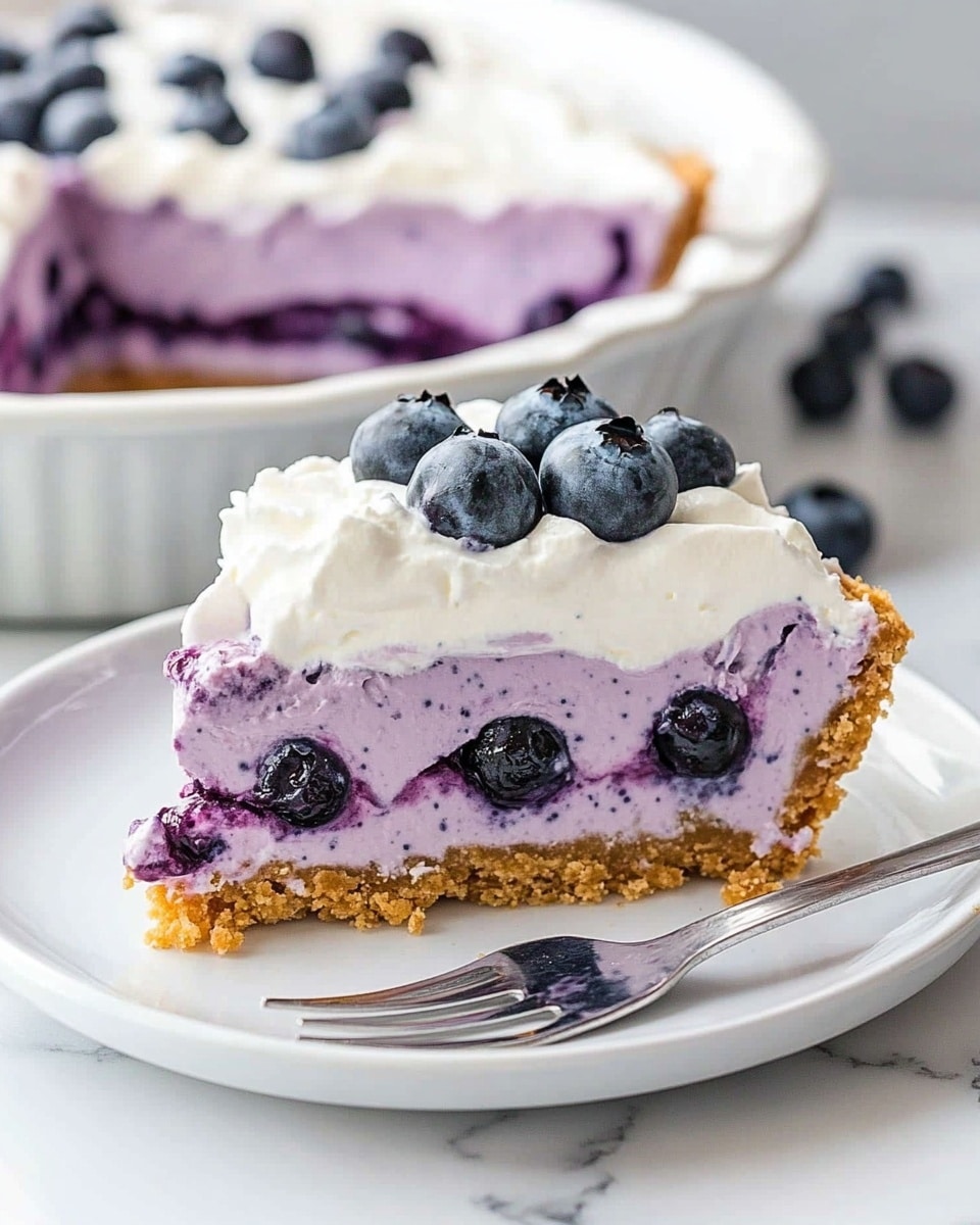 No Bake Blueberry Cream Pie, blueberry dessert, easy summer pie, no bake fruit dessert, creamy blueberry pie The image shows a slice of three-layer blueberry dessert on a white plate with a silver fork beside it. The bottom layer is a crumbly golden-brown crust. The middle layer is thick and creamy with a purple hue mixed with whole blueberries, looking soft and rich. The top layer is smooth white whipped cream, topped with a single fresh blueberry. In the background, there is a white dish with more of the dessert, placed on a white marbled surface. The