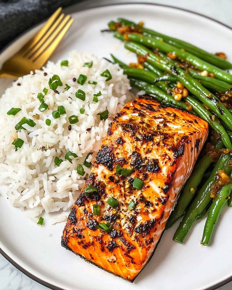 Cajun Honey Butter Salmon, Cajun Salmon Recipe, Honey Butter Salmon, Spicy Sweet Salmon, Easy Salmon Dinner A white plate holds three main layers: a portion of white rice sprinkled with small black and green herbs on the left, a serving of cooked green beans with minced garlic pieces in the middle, and a thick grilled salmon fillet with a dark brown, slightly charred crust on the right. The salmon fillet is partially sliced to show its tender, light pink inside and is garnished with finely chopped green herbs. A gold fork with a black handle rests on the plate near the salmon. The plate sits on a white marbled textured surface.
