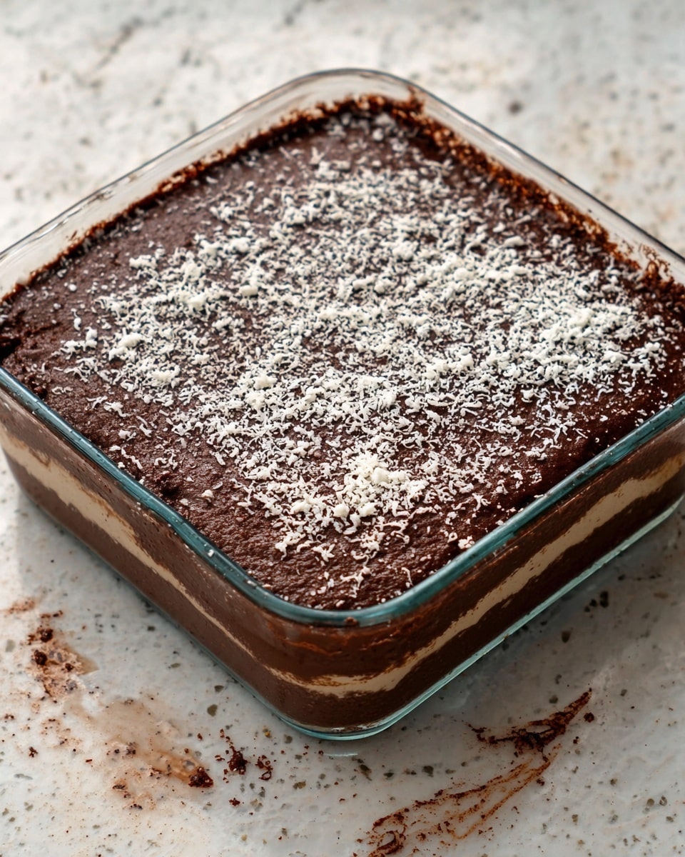 A square clear glass baking dish holds a chocolate dessert with three visible layers: the bottom layer is dark brown and dense, the middle layer is a slightly lighter chocolate brown with a soft texture, and the top layer is dark chocolate covered evenly with a scattering of fine white shavings. The dish sits on a white marbled surface, showing some small spills of chocolate around the edges. photo taken with an iphone --ar 4:5 --v 7
