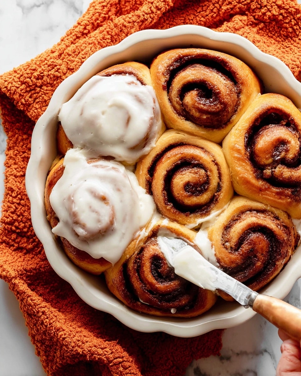 A close-up view of a single cinnamon roll on a white plate showing at least three thick layers of soft, orange-brown dough swirled with dark brown cinnamon filling. The top is covered with a thick, creamy white glaze that drips down the sides, adding a smooth texture contrast to the fluffy dough. The background features a blurred second cinnamon roll and a clean white marbled surface under the plate. The warm tones of the cinnamon roll stand out against the white plate and surface, creating a cozy and inviting look. photo taken with an iphone --ar 4:5 --v 7