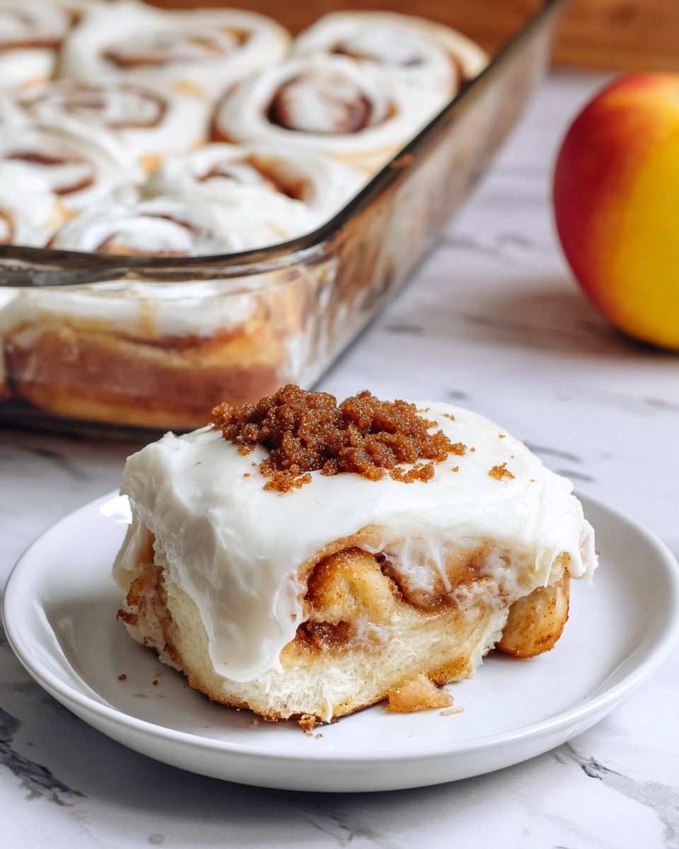 A close-up view of a cinnamon roll on a white plate showing three main layers: the bottom layer is a thick, soft, light beige dough, the middle layer is a golden brown swirl of cinnamon and apple pieces giving a sticky texture, and the top layer is a smooth, thick white frosting covering the roll with a small pile of crumbly dark brown streusel on top. The plate sits on a wooden surface, and in the background, there's a glass baking dish filled with more cinnamon rolls covered in the same white frosting. Photo taken with an iphone --ar 4:5 --v 7
