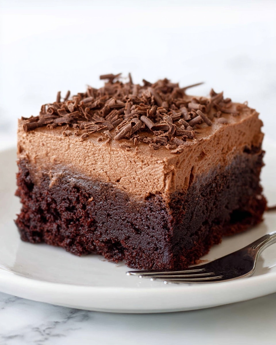 A square piece of chocolate cake with two distinct layers sits on a white plate over a white marbled surface. The bottom layer is dark brown, dense, and slightly moist with a rough texture. The top layer is thick, creamy, and lighter brown chocolate frosting spread evenly, topped with small, curly chocolate shavings scattered across its surface. A silver fork lies near the bottom right edge of the plate. Photo taken with an iphone --ar 4:5 --v 7