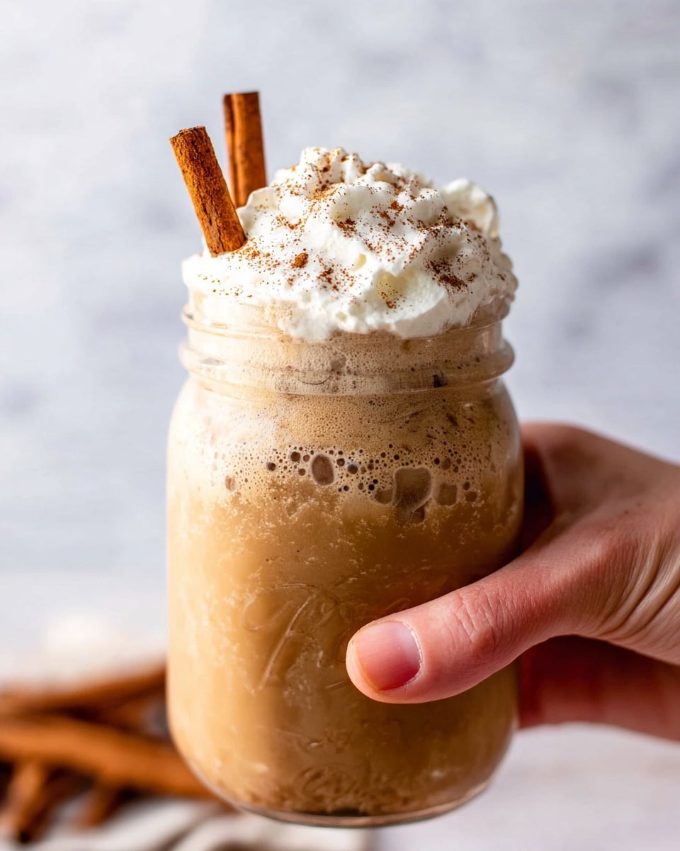 A clear glass jar filled with a creamy light brown drink fills most of the jar with some ice visible inside. On top, there is a thick layer of white whipped cream sprinkled lightly with brown powder. Two whole cinnamon sticks are placed upright inside the jar, leaning slightly to one side. The jar sits on a light wooden cutting board with a rustic texture, and there is a light dusting of cinnamon powder on the board and the white marbled surface around it. In the background, blurred cinnamon sticks and star anise add depth to the scene. photo taken with an iphone --ar 4:5 --v 7