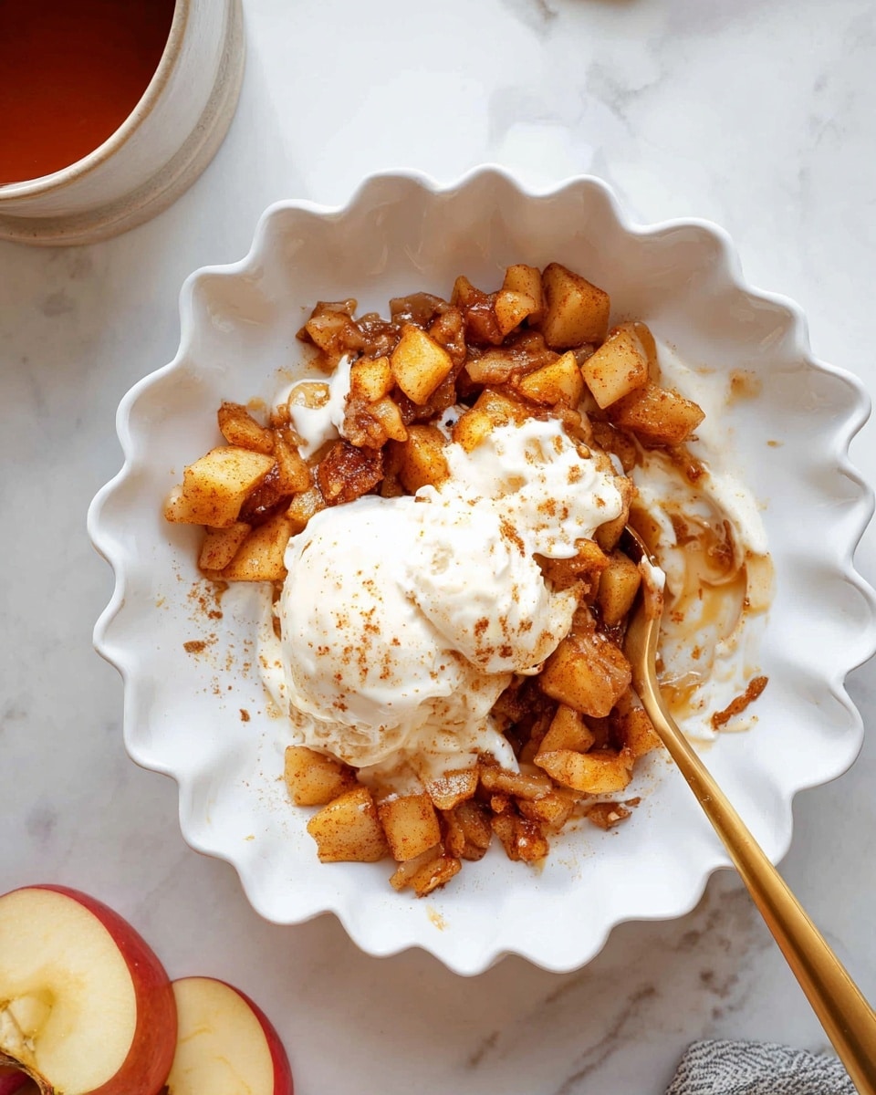 A white scalloped bowl holds a warm dessert of two main layers: the bottom layer is a mix of small cooked apple chunks with a golden-brown color and a slightly caramelized texture, scattered unevenly. The top layer is soft, creamy vanilla ice cream melting over the warm apples, creating a smooth, white contrast to the rich brown fruit. A golden spoon rests inside the bowl, partially covered by the apple pieces and ice cream. The bowl sits on a white marbled surface with apple slices and a cup of tea nearby. photo taken with an iphone --ar 4:5 --v 7