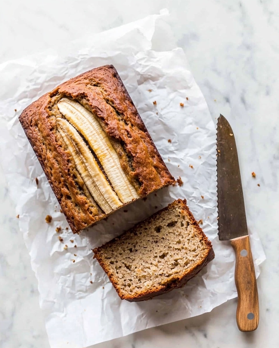 A loaf of banana bread is on white parchment paper over a white marbled surface. The bread is partly sliced into five thick pieces with a visible crust that is dark brown and textured with a swirl pattern of bananas on top. One slice is fully separated and lies flat, showing a soft, light brown crumb with small holes and bits of banana inside. A wooden-handled knife rests beside the slice on the right. The overall setting is bright with natural light. photo taken with an iphone --ar 4:5 --v 6.1